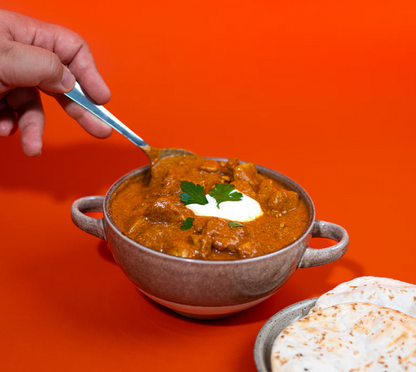 A hand holding a spoon above a bowl of creamy butter chicken with a dollop of yogurt, served with flatbreads.