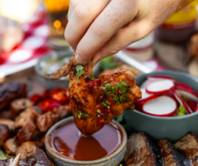 Hand dipping chicken wing into sauce surrounded by assorted grilled meats and fresh vegetables.